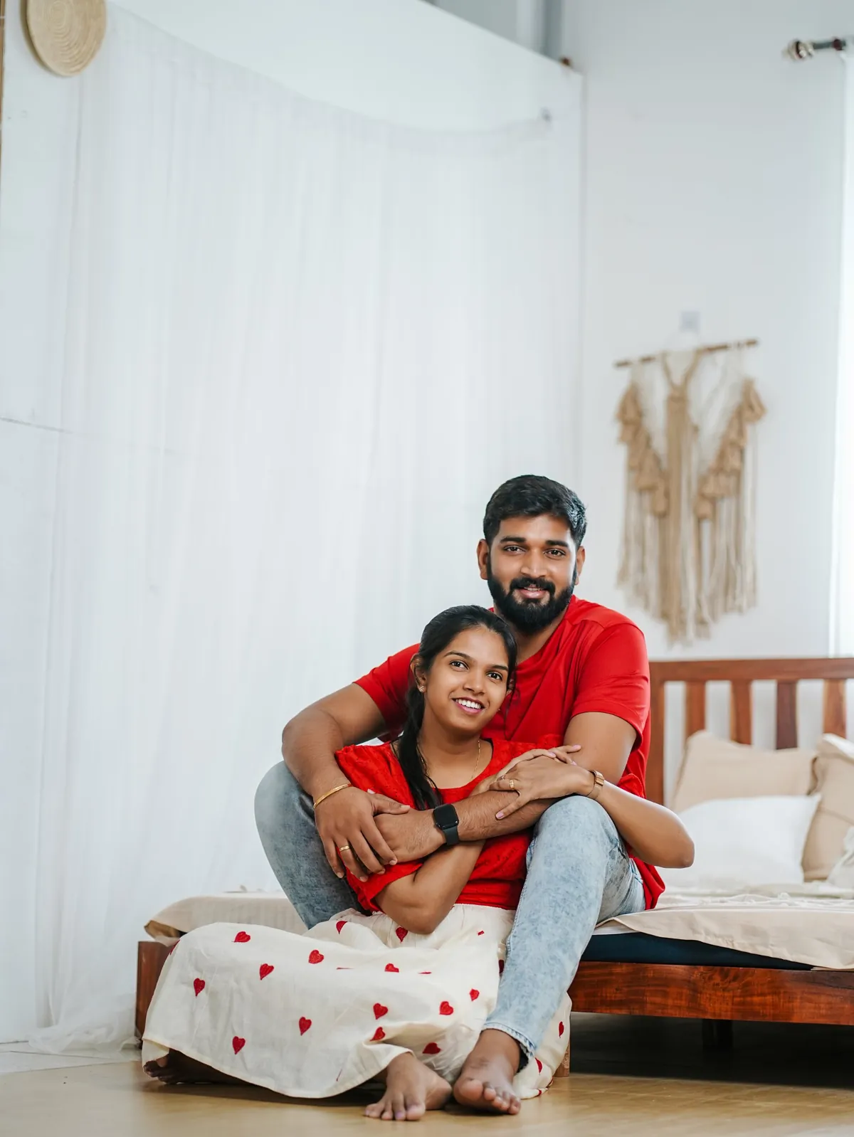 A couple in red sit by a low wooden bed in a bright white room; he wraps his arms around her from behind and both smile at the camera.