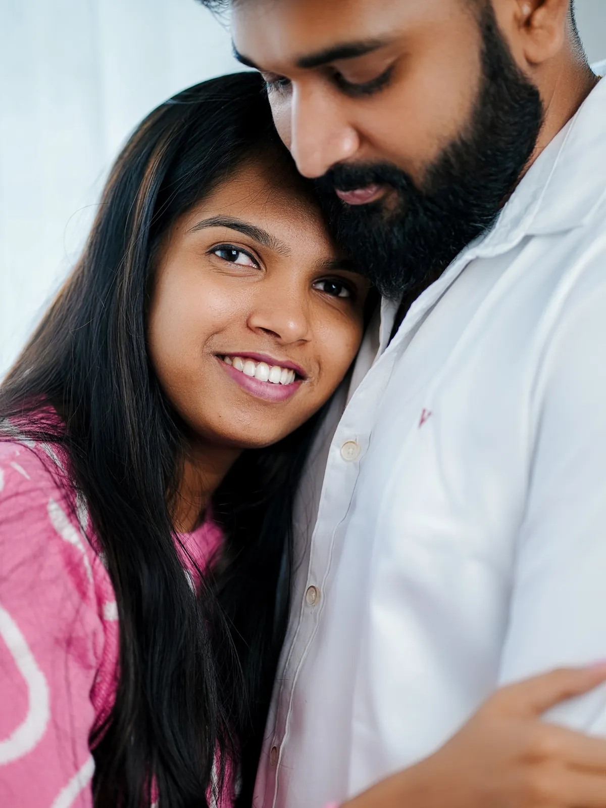 Close portrait of a woman in pink smiling at the camera with her head against the chest of her partner, who looks down at her.