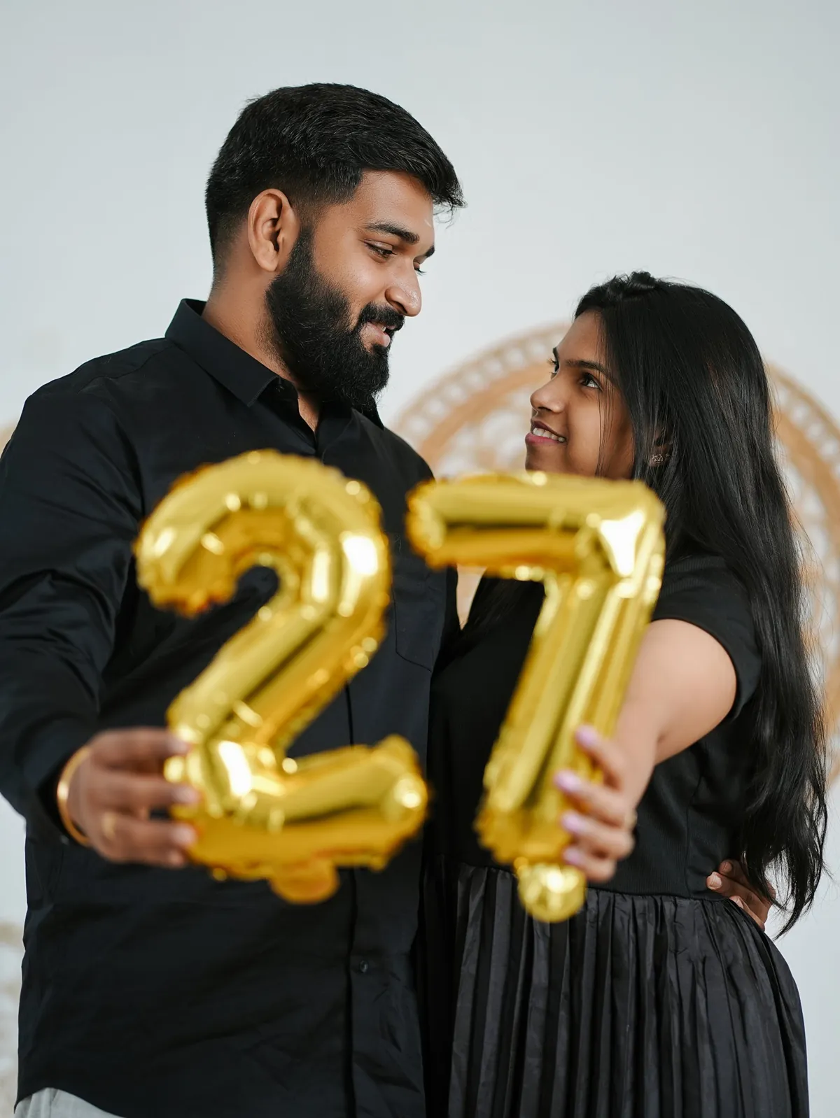 A couple in black look at each other while holding gold foil balloons shaped as the number 27 towards the camera.
