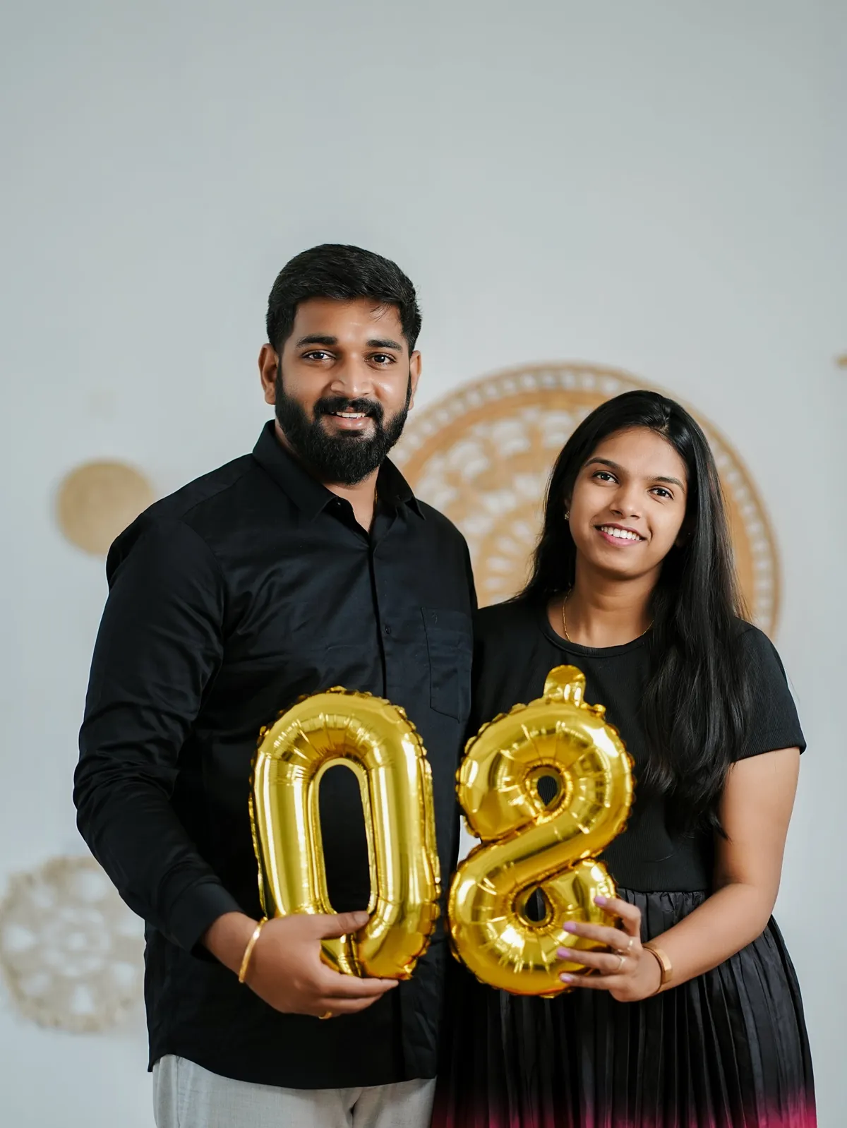 The same couple in black smile at the camera, holding gold foil balloons shaped as 08.