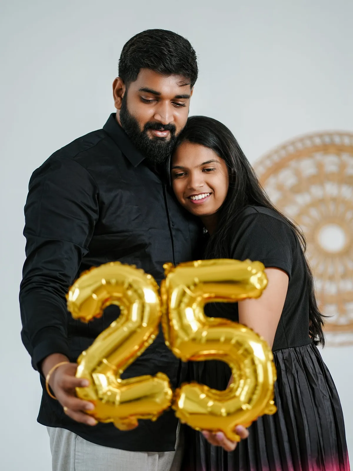 The couple in black embrace and look down at gold foil balloons shaped as the number 25.