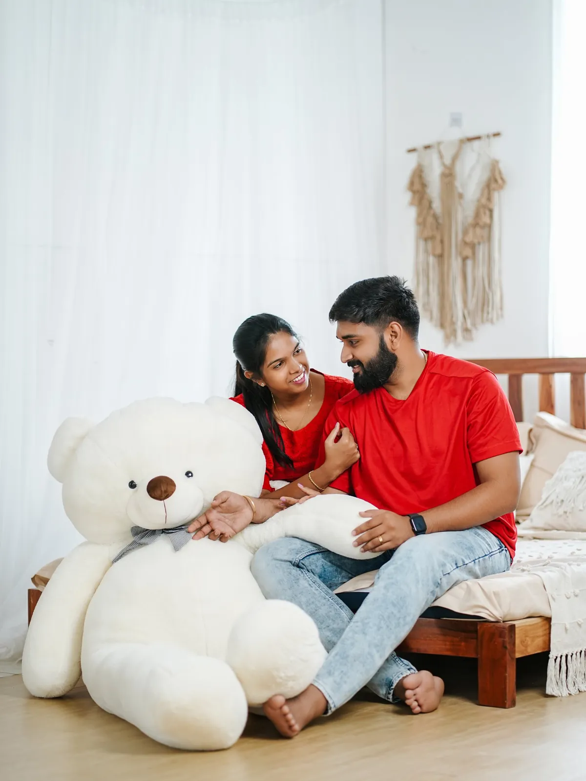 A couple in red sit on a low bed smiling at each other beside a giant white teddy bear.