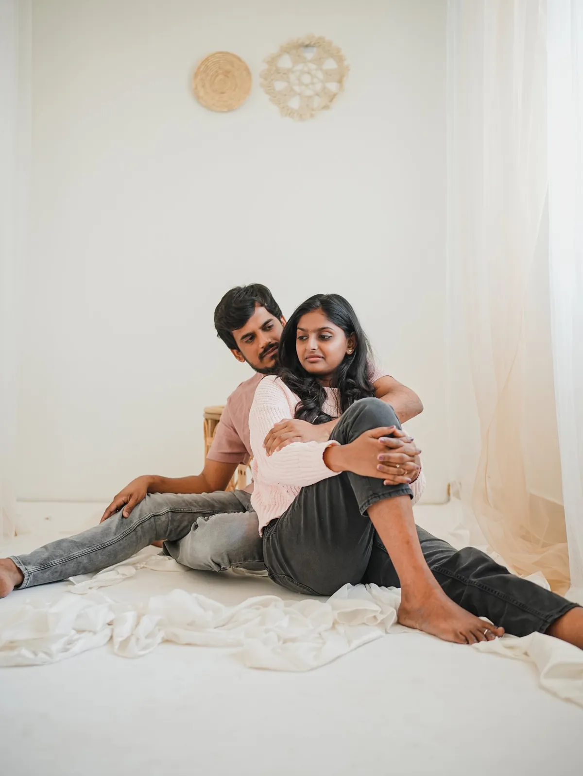 A couple in soft pink tops sit on a white studio floor among draped fabric; he holds her from behind as she looks away.