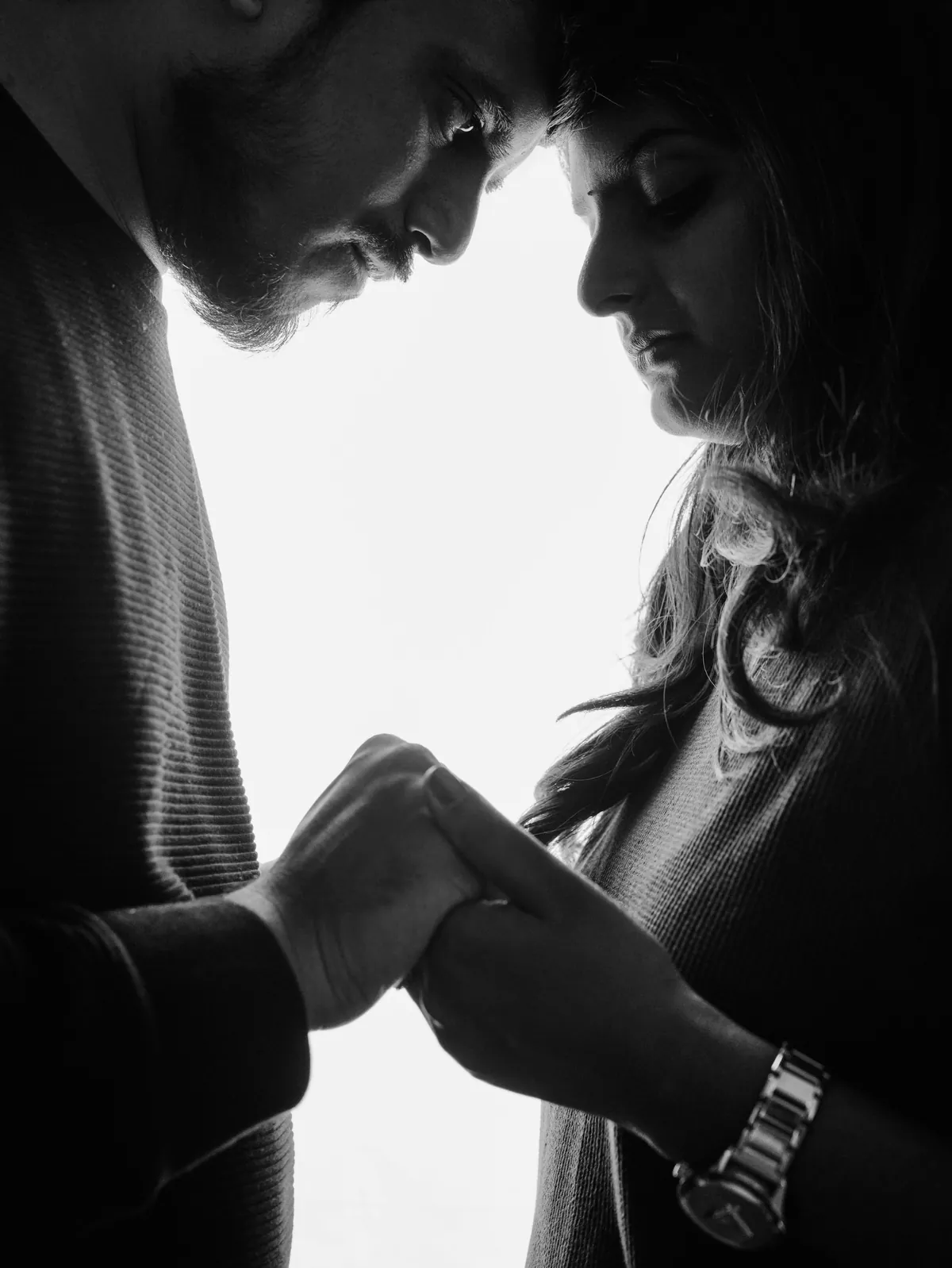 Backlit black and white close-up of a couple with foreheads touching and hands clasped between them.