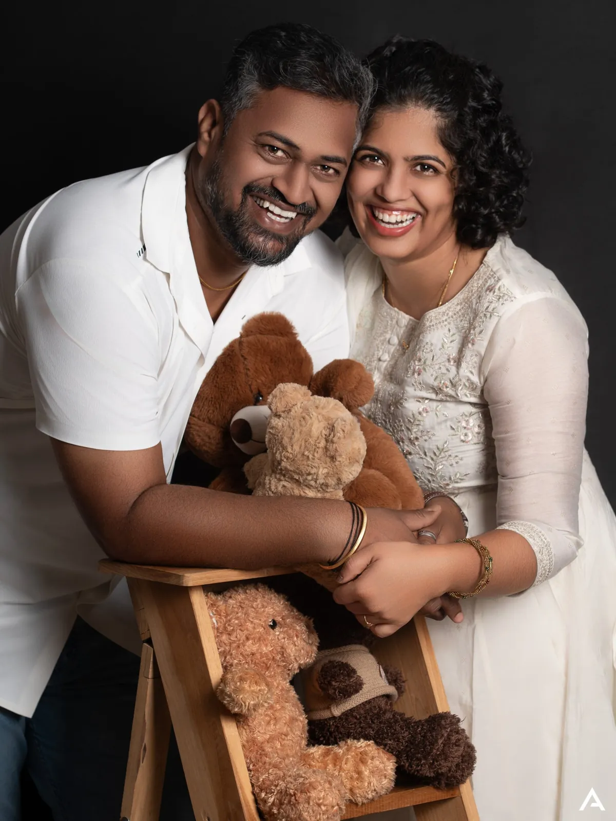 A laughing couple in white lean together over a wooden ladder stacked with teddy bears, against a black backdrop.
