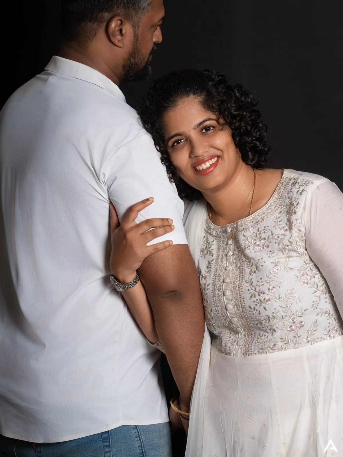 A woman in an embroidered white dress smiles at the camera, resting her head on the shoulder of her partner, who faces away.