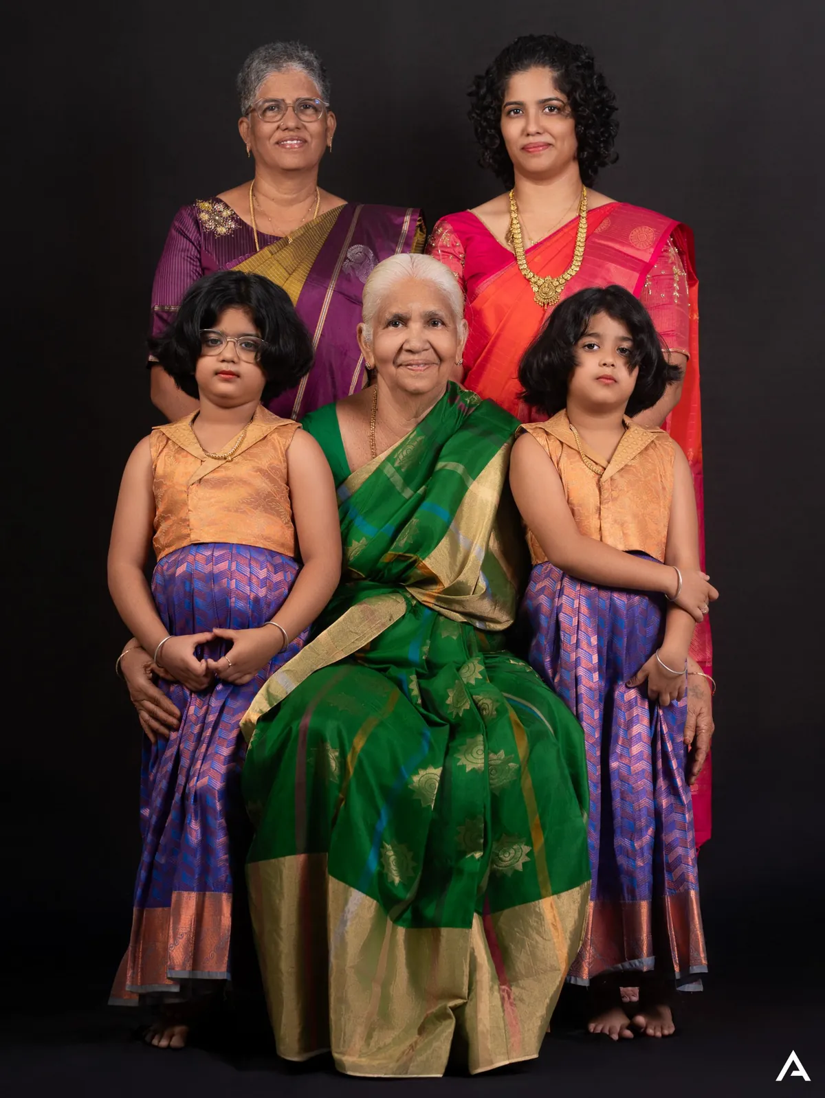 Formal portrait of five women and girls of one family in bright silk saris and pavadai; the eldest is seated at the centre in green against a black backdrop.