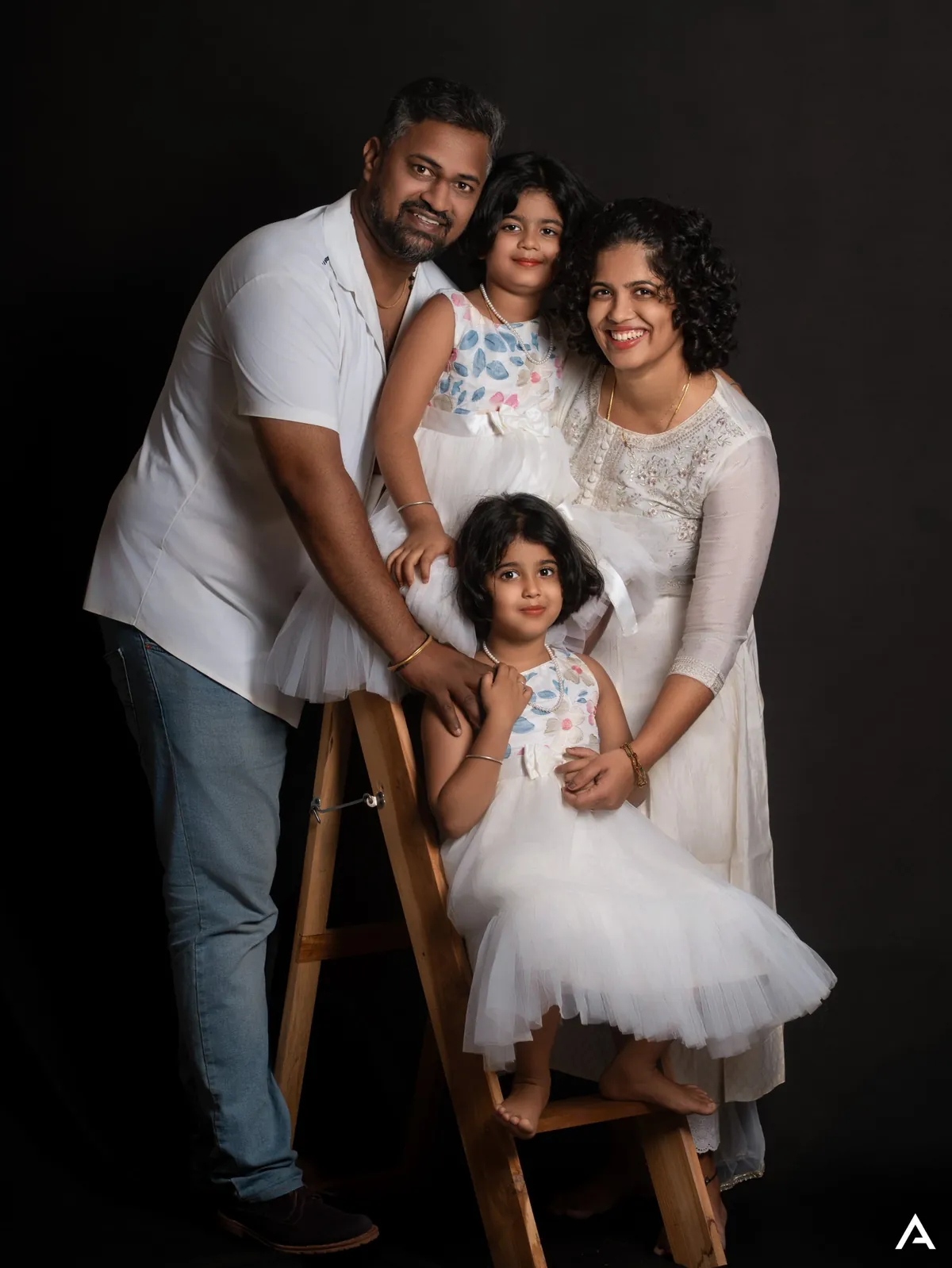 Parents and two young daughters in white pose around a wooden ladder against a black backdrop, all smiling.