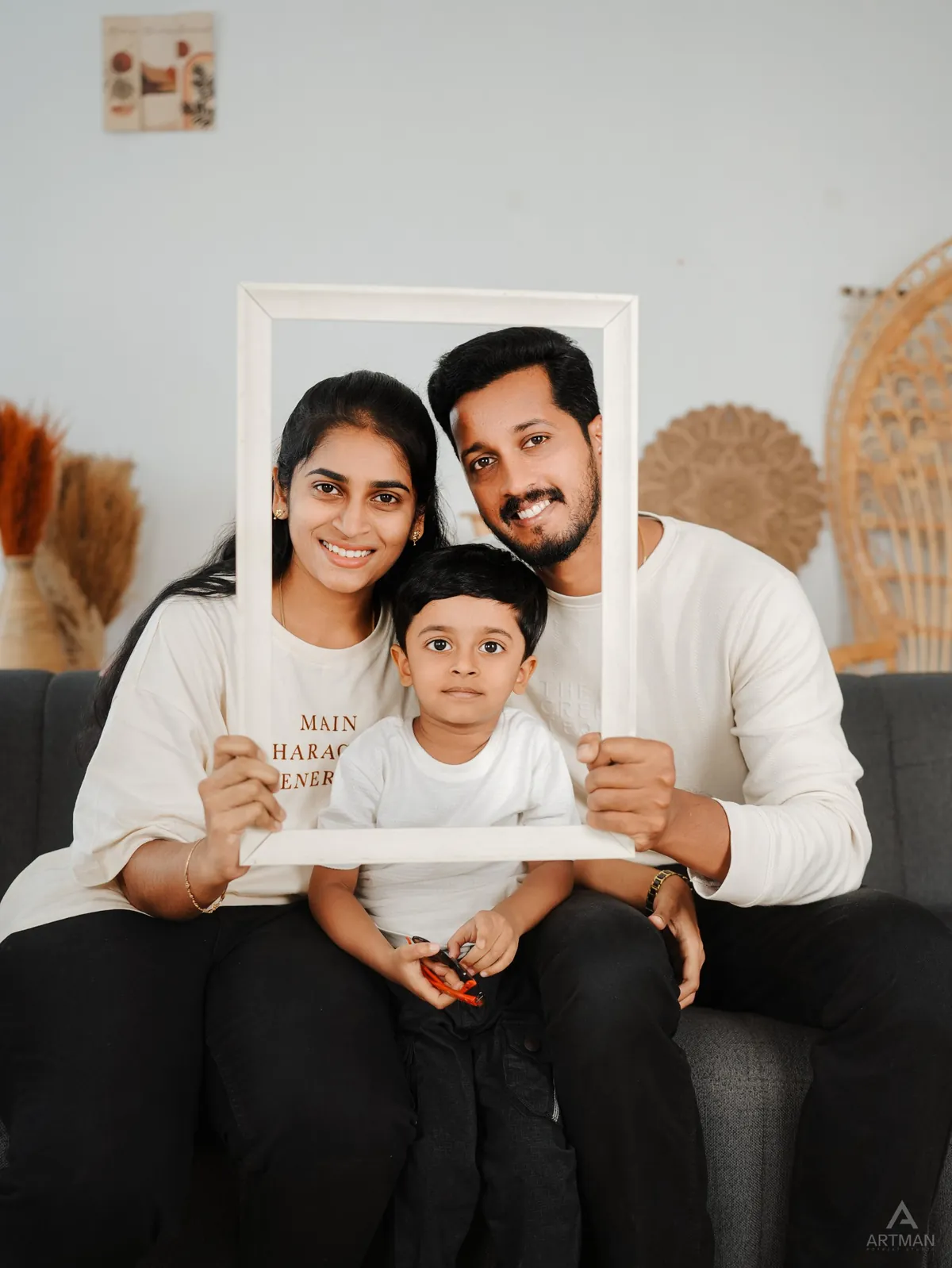 A couple and their young son sit on a sofa, holding an empty white picture frame around their faces.