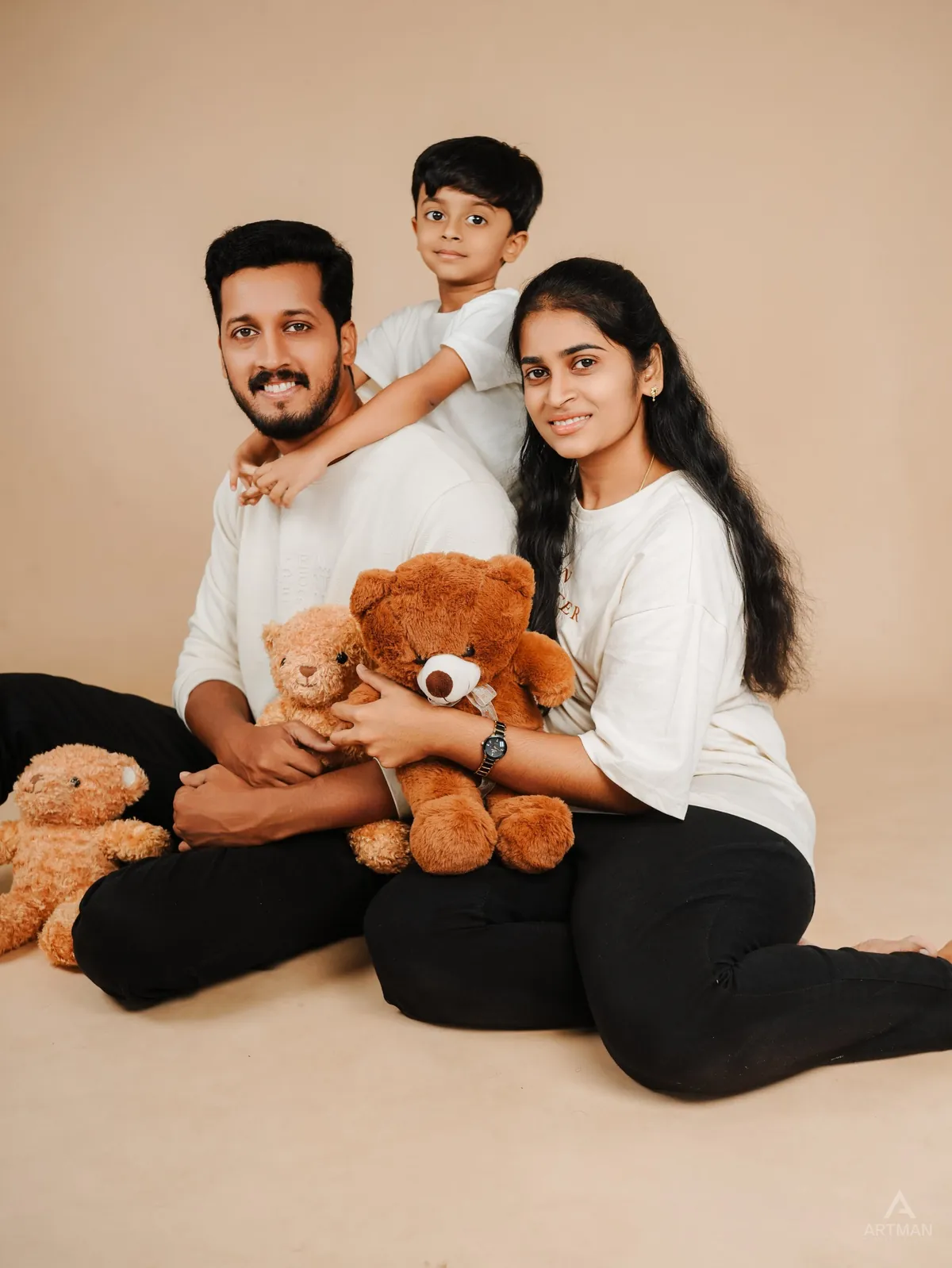 Parents sit on the studio floor with teddy bears while their son leans over his father's shoulders, against a beige backdrop.