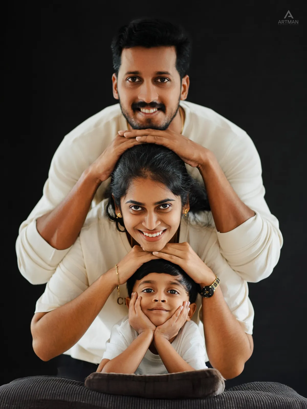 A father, mother and young son stack their chins on each other's heads, smiling, against a black backdrop.