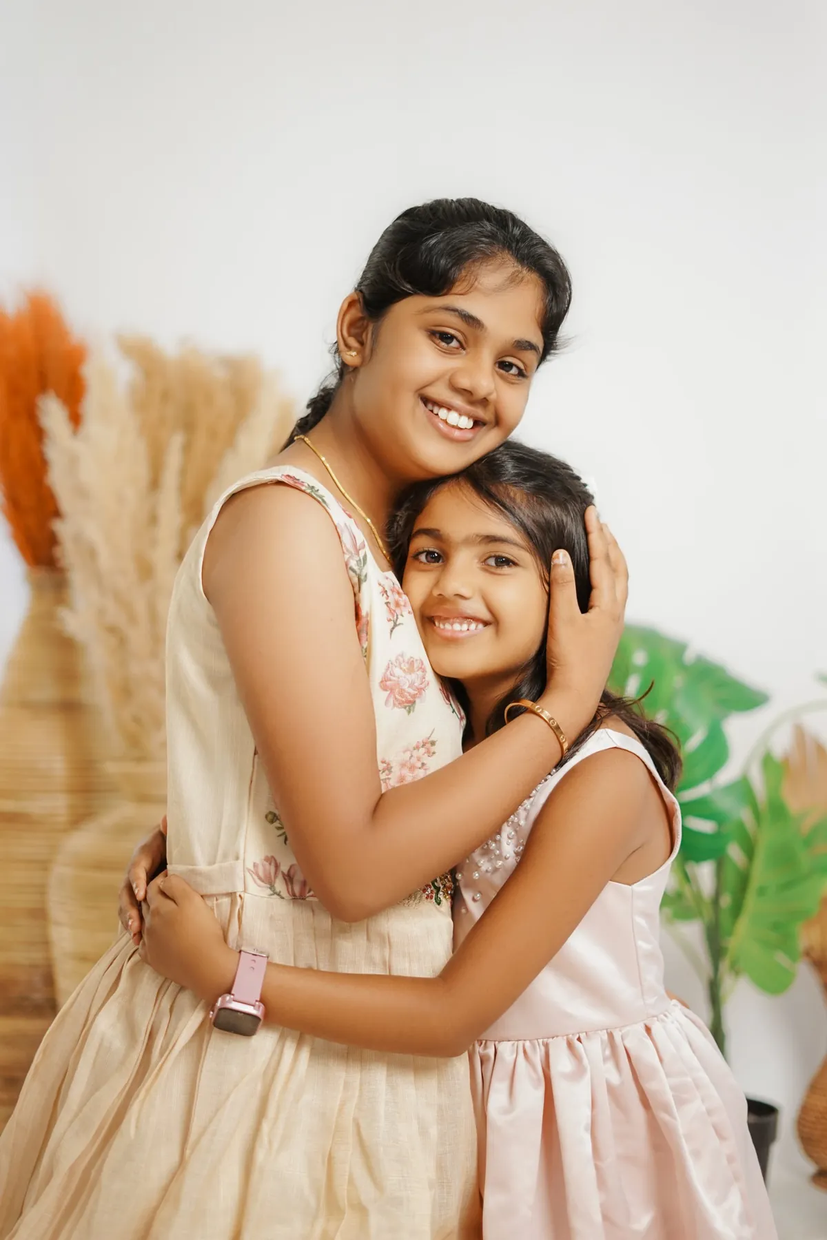 Two sisters hug and smile at the camera; the elder cradles the younger one's head, with pampas grass and green leaves behind.