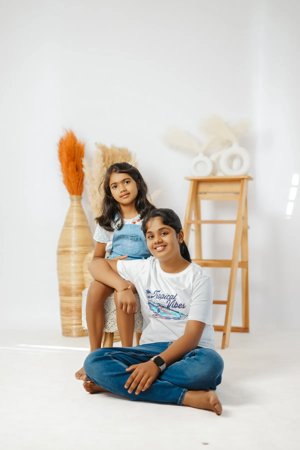 Two sisters in casual clothes pose on a white set; the elder sits cross-legged on the floor and the younger sits on a stool behind her.