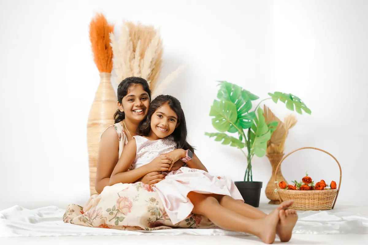 Two sisters sit laughing on a white sheet, the younger leaning back in the elder's lap, beside pampas grass, a leafy plant and a basket of roses.
