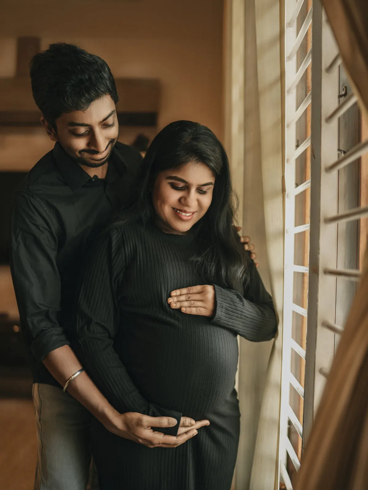 A couple in black stand by a sunlit window; he holds her from behind and both look down at her bump, smiling.