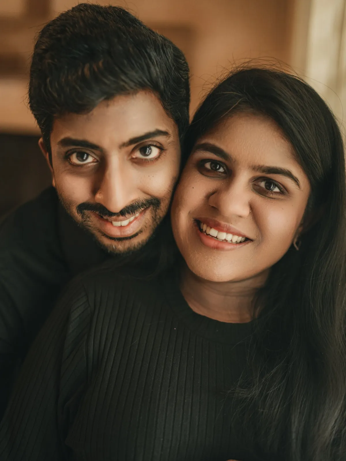 Close portrait of a smiling couple in black, cheeks touching, looking into the camera.
