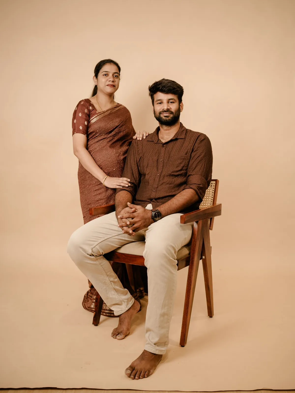 A woman in a brown silk sari stands with her hand on the shoulder of her partner, who sits in a cane-back chair against a beige backdrop.