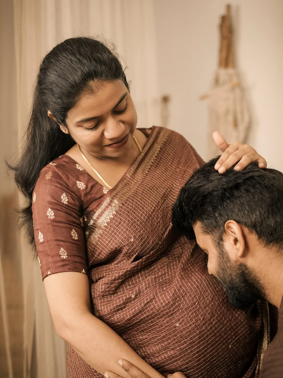 A woman in a brown silk sari smiles down as her partner bows his head to her bump; her hand rests on his hair.