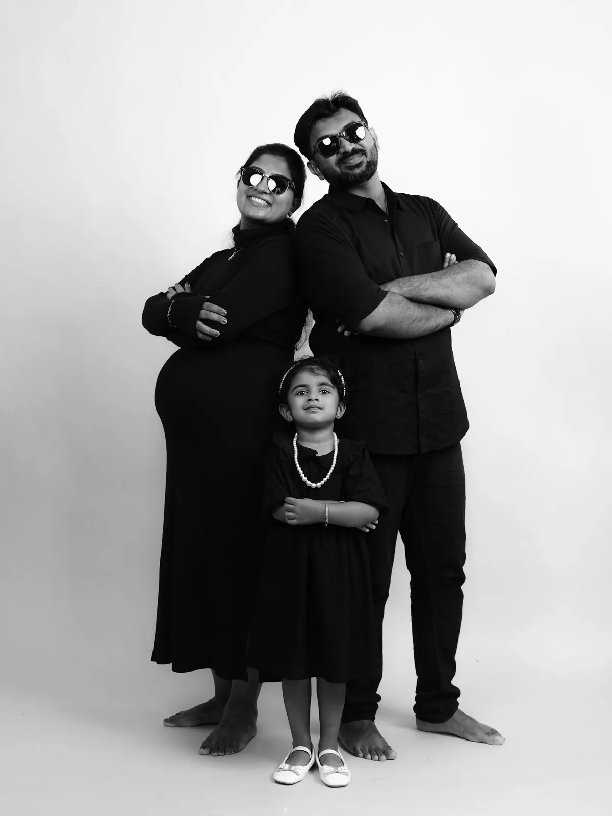 Black and white studio portrait of expectant parents standing back to back in sunglasses with arms folded, their young daughter copying the pose in front.