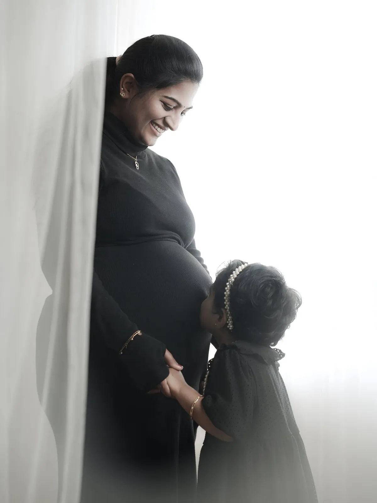 A mother in black leans against a white curtain, laughing down at her young daughter, who kisses the bump.