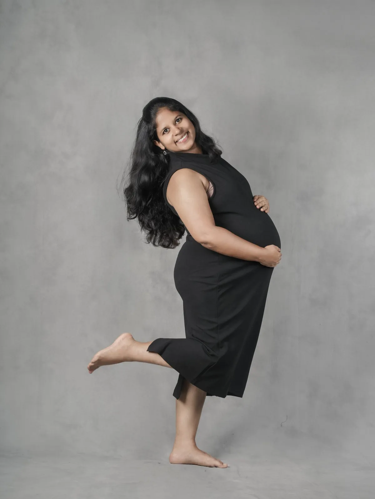 A woman in a black dress laughs towards the camera, one foot kicked up behind her, against a grey painted backdrop.
