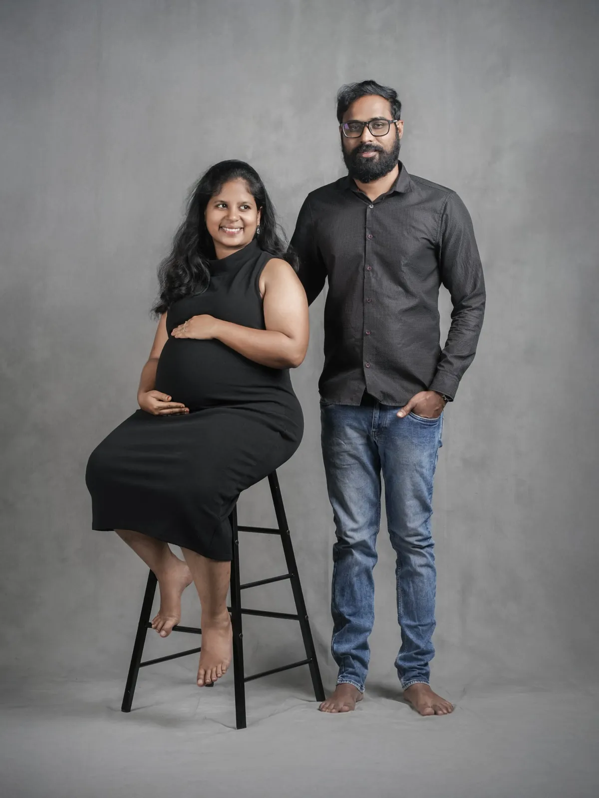 A woman in a black dress sits on a tall stool holding her bump and smiling away from camera; her partner stands beside her against a grey backdrop.