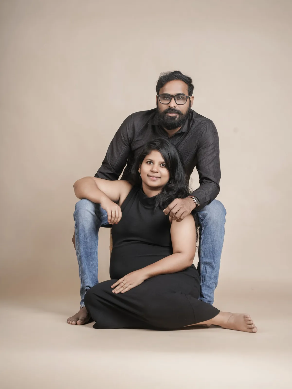 A woman in black sits on the studio floor leaning against her partner, who sits behind her with a hand on her shoulder, against a beige backdrop.