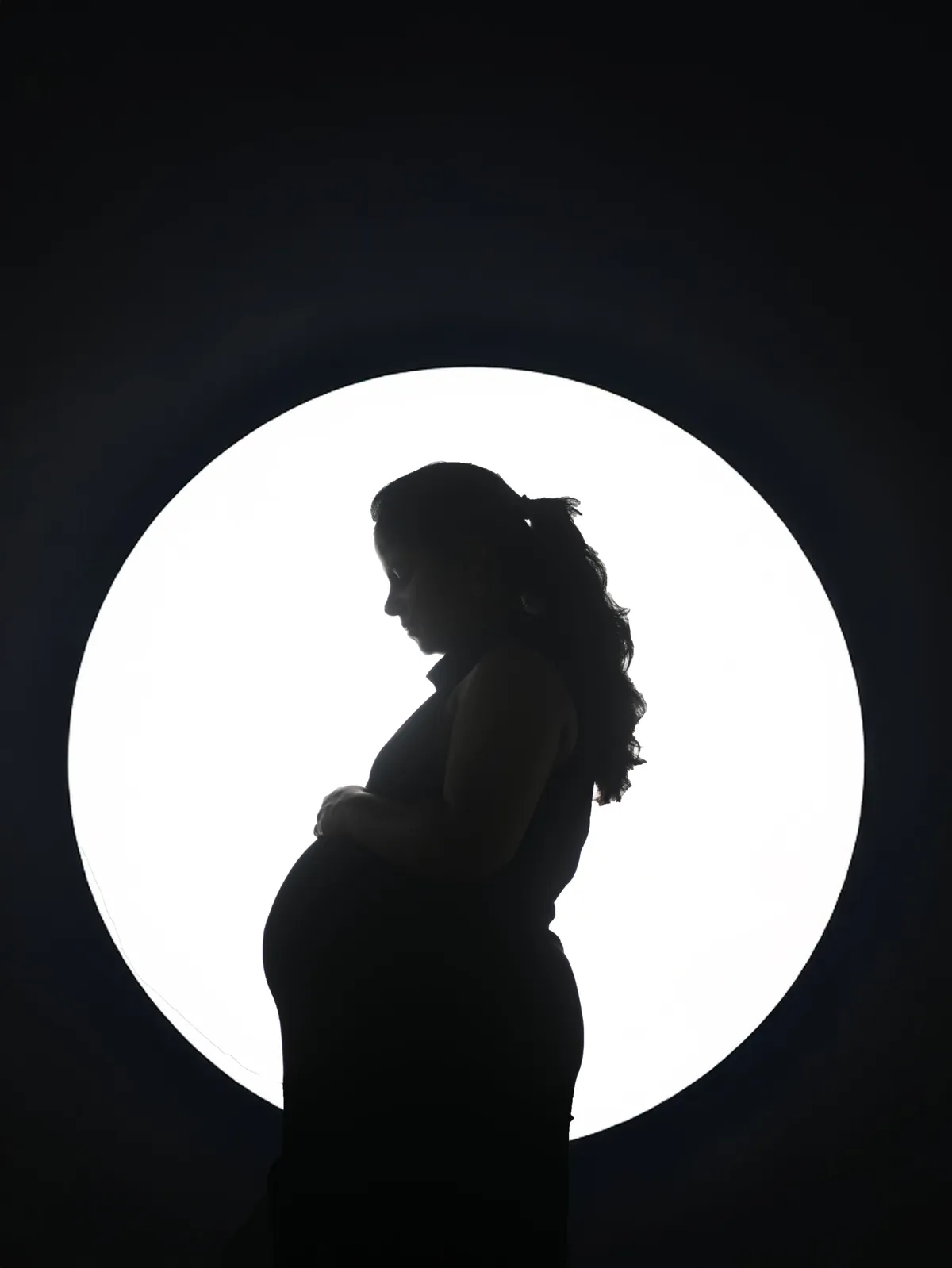 Silhouette of a pregnant woman in profile, head bowed and hand on her bump, against a glowing white disc on a black background.