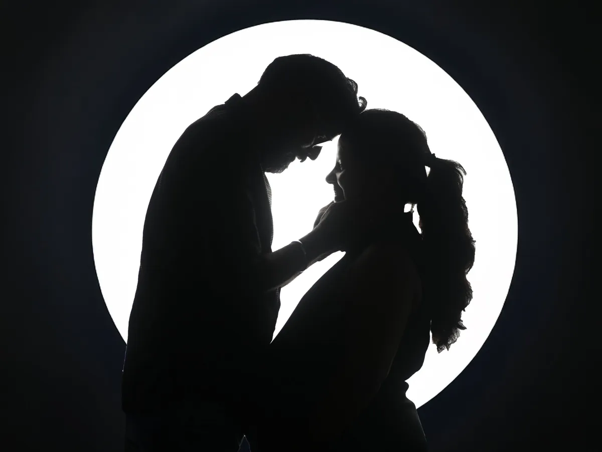 Silhouette of a couple against a glowing white disc; he leans in to touch his forehead to hers and cups her face as she smiles.