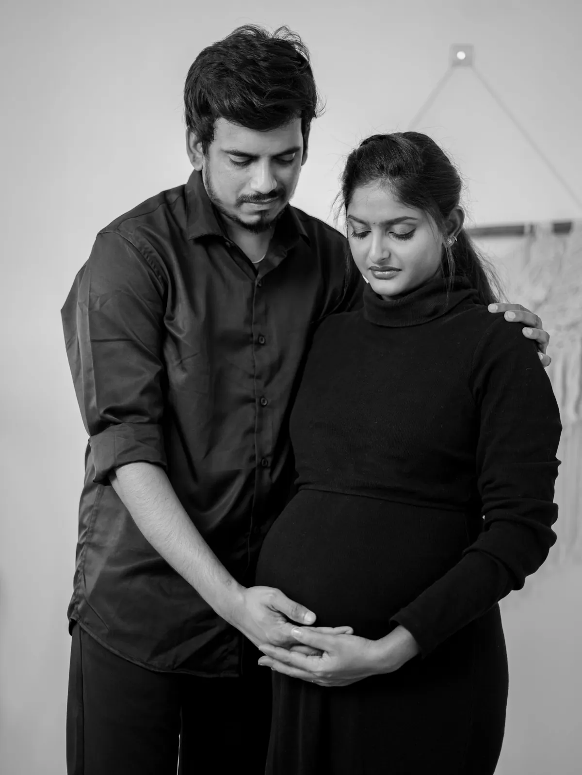Black and white portrait of expectant parents in black looking down, his hand on her shoulder and their hands joined under her bump.