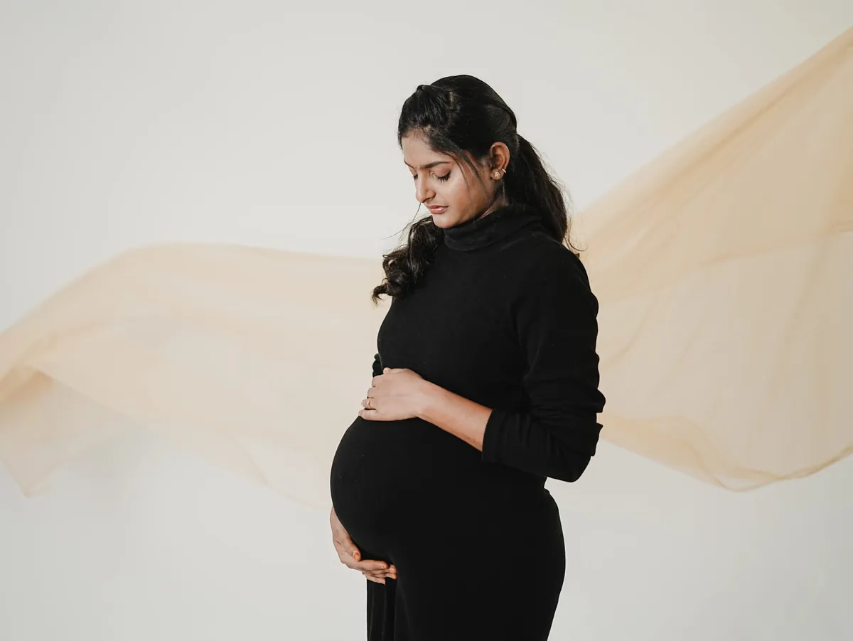 A pregnant woman in a black dress looks down and cradles her bump while a length of sheer beige fabric floats across the white backdrop behind her.