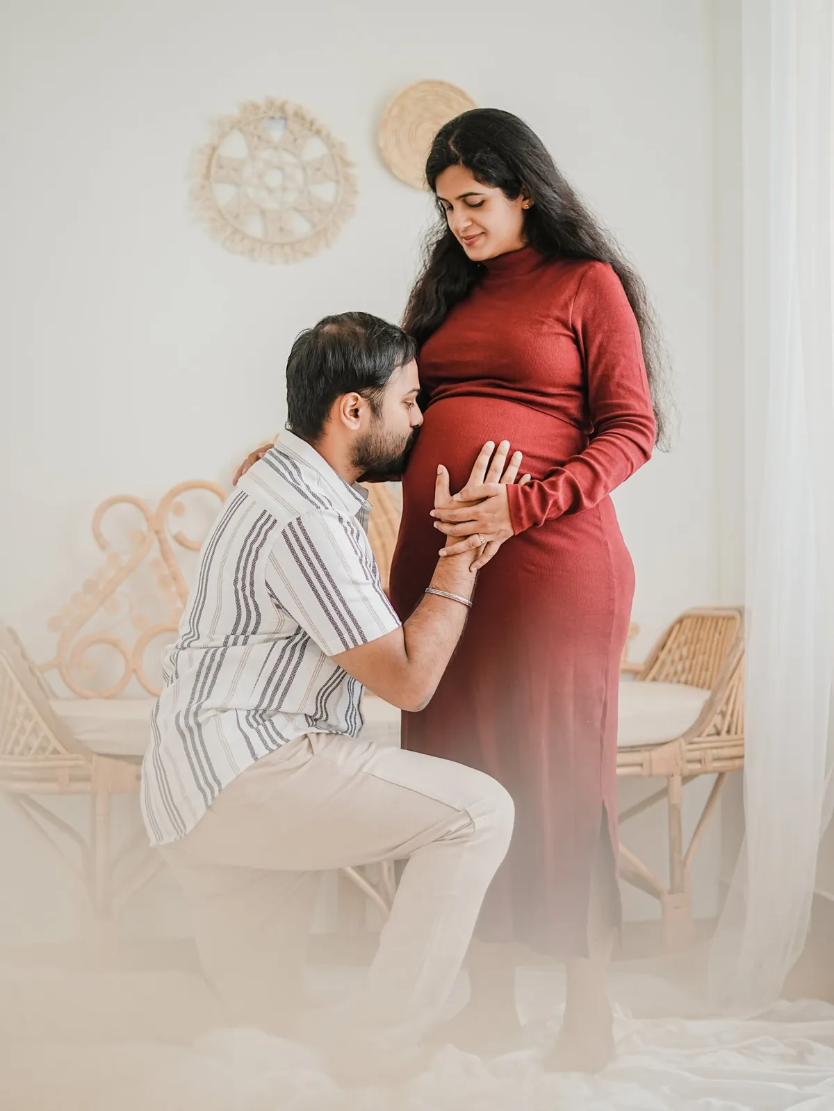 A man kneels to kiss the bump of his partner, who stands in a rust dress and looks down at him, in a white room with rattan furniture.