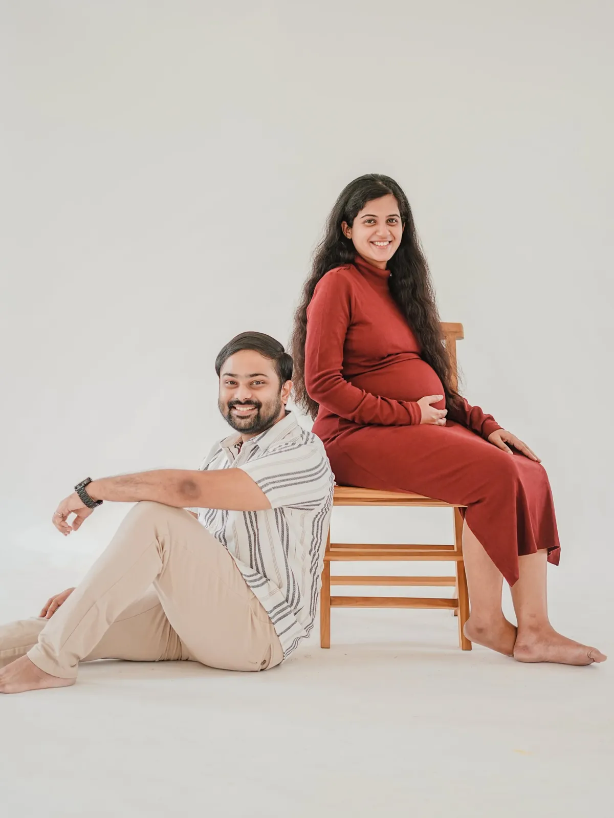 A pregnant woman in a rust dress sits on a wooden stool smiling at the camera while her partner sits on the floor leaning against it, on a white backdrop.