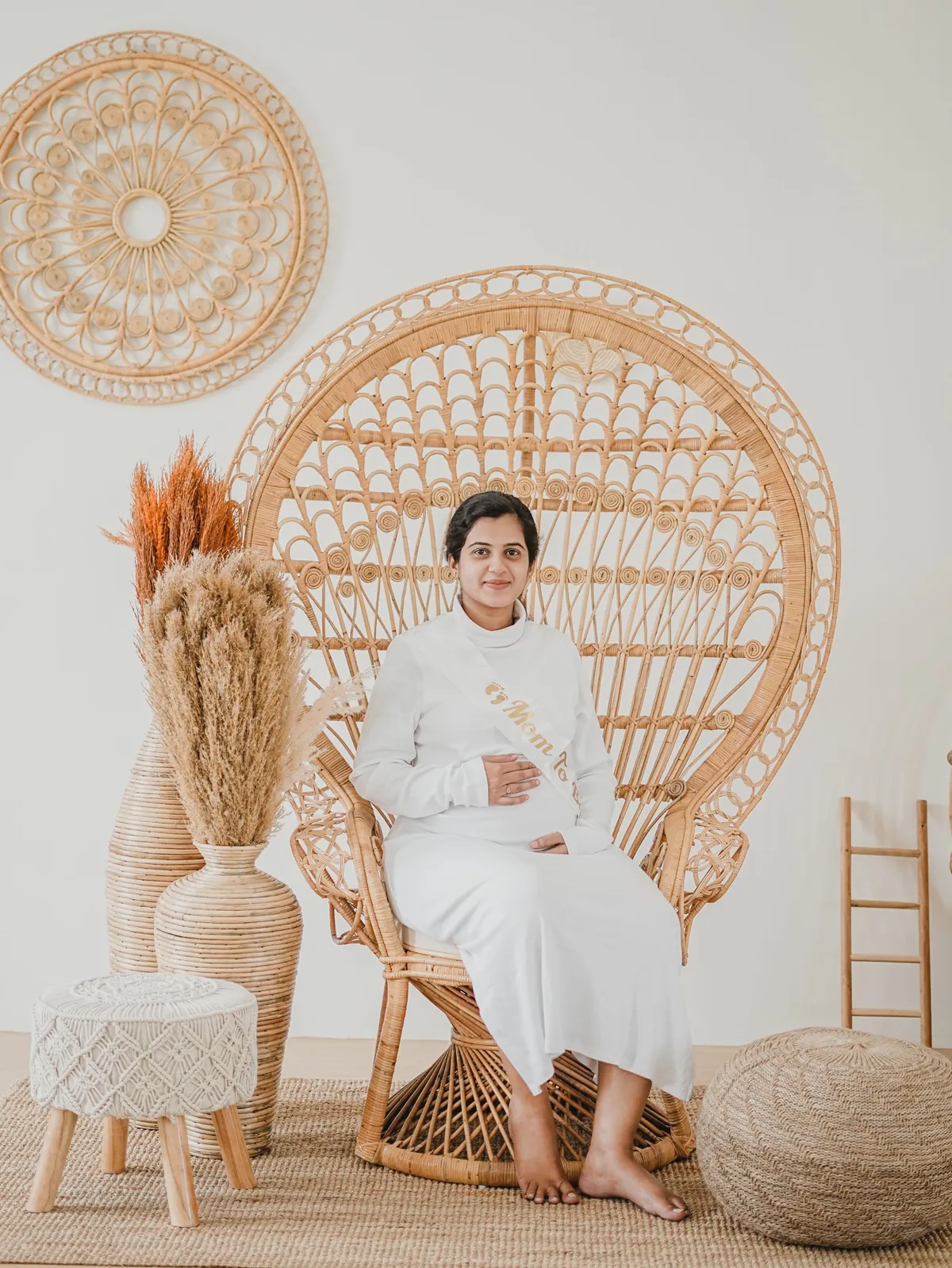 A pregnant woman in white sits in a tall rattan peacock chair wearing a mum-to-be sash, surrounded by pampas grass and woven decor.