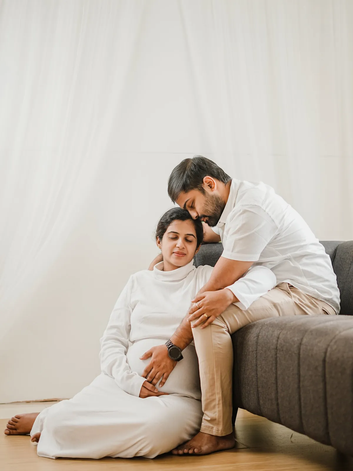 A pregnant woman in white sits on the floor with her eyes closed, leaning against her partner, who sits on a sofa and kisses the top of her head.