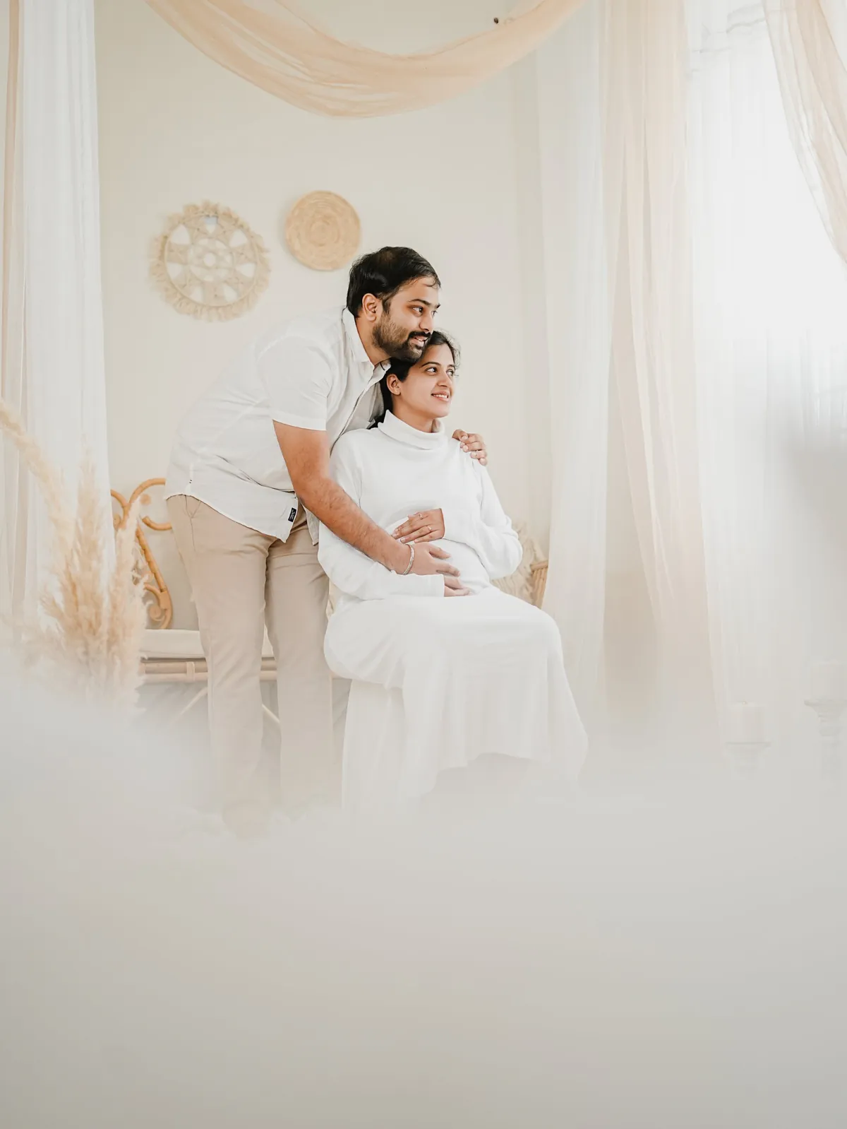 A couple in white look towards a window, his hands on her shoulder and bump, photographed through soft white fabric in the foreground.