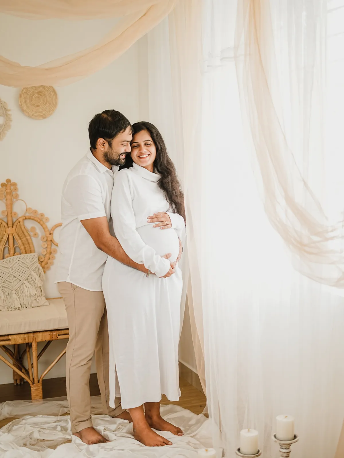 A couple in white stand at a bright curtained window; he holds her from behind and both smile, with rattan furniture and candles around them.