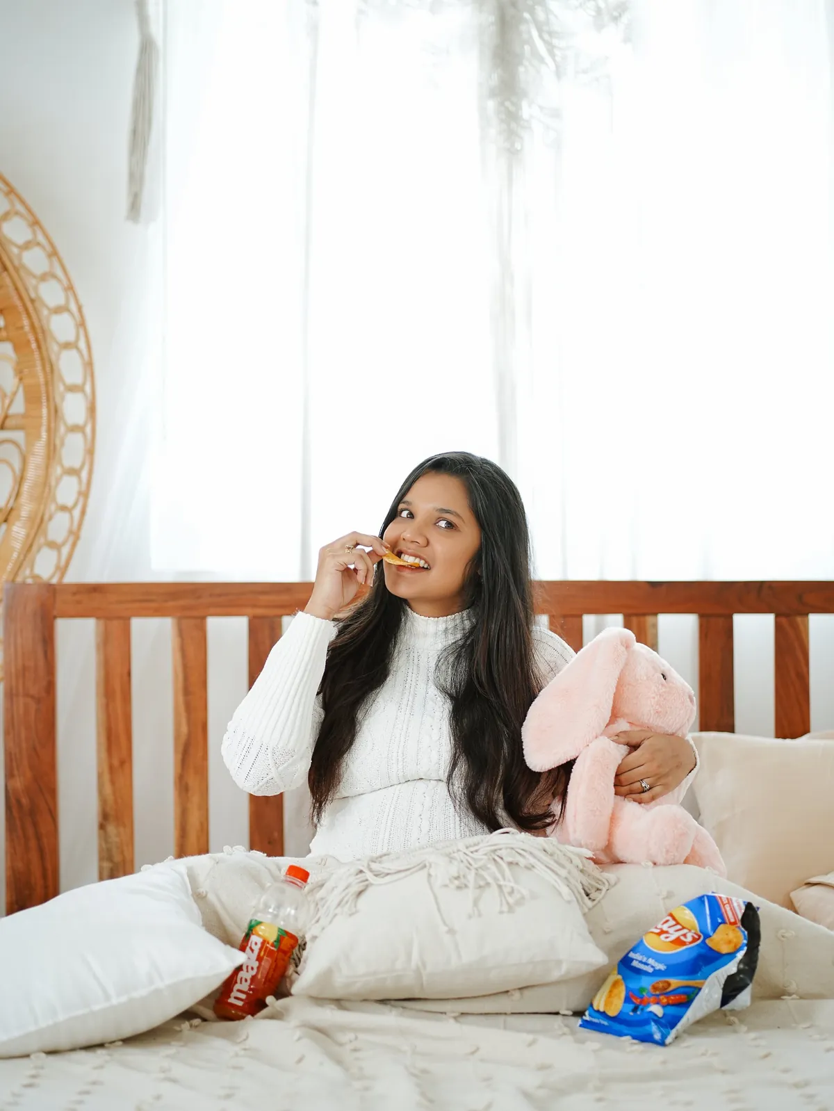 A mother-to-be in a white knit dress sits on a bed eating a crisp and hugging a pink soft-toy rabbit, with a drink and a snack packet beside her.