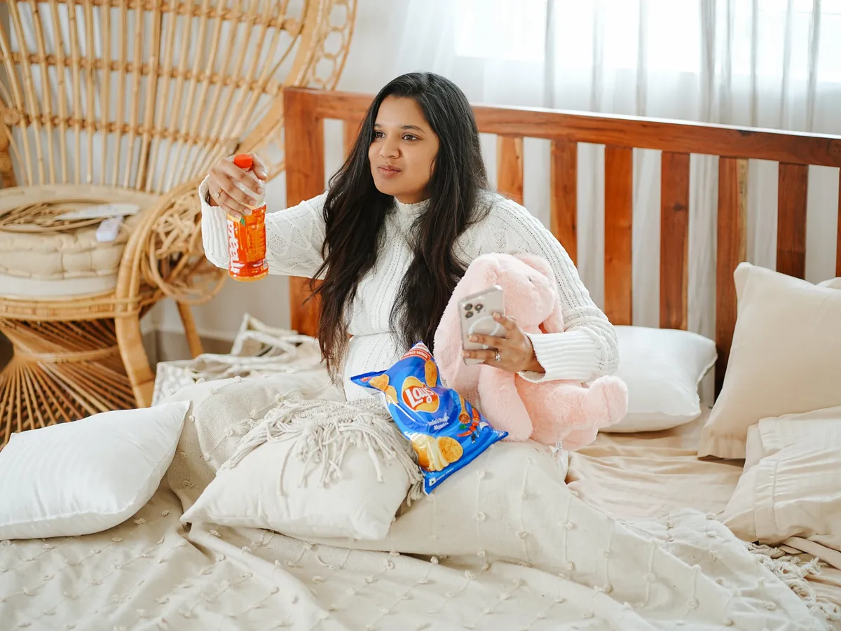 A mother-to-be sits on a bed among cushions holding a drink bottle in one hand and a phone and a pink soft-toy rabbit in the other.