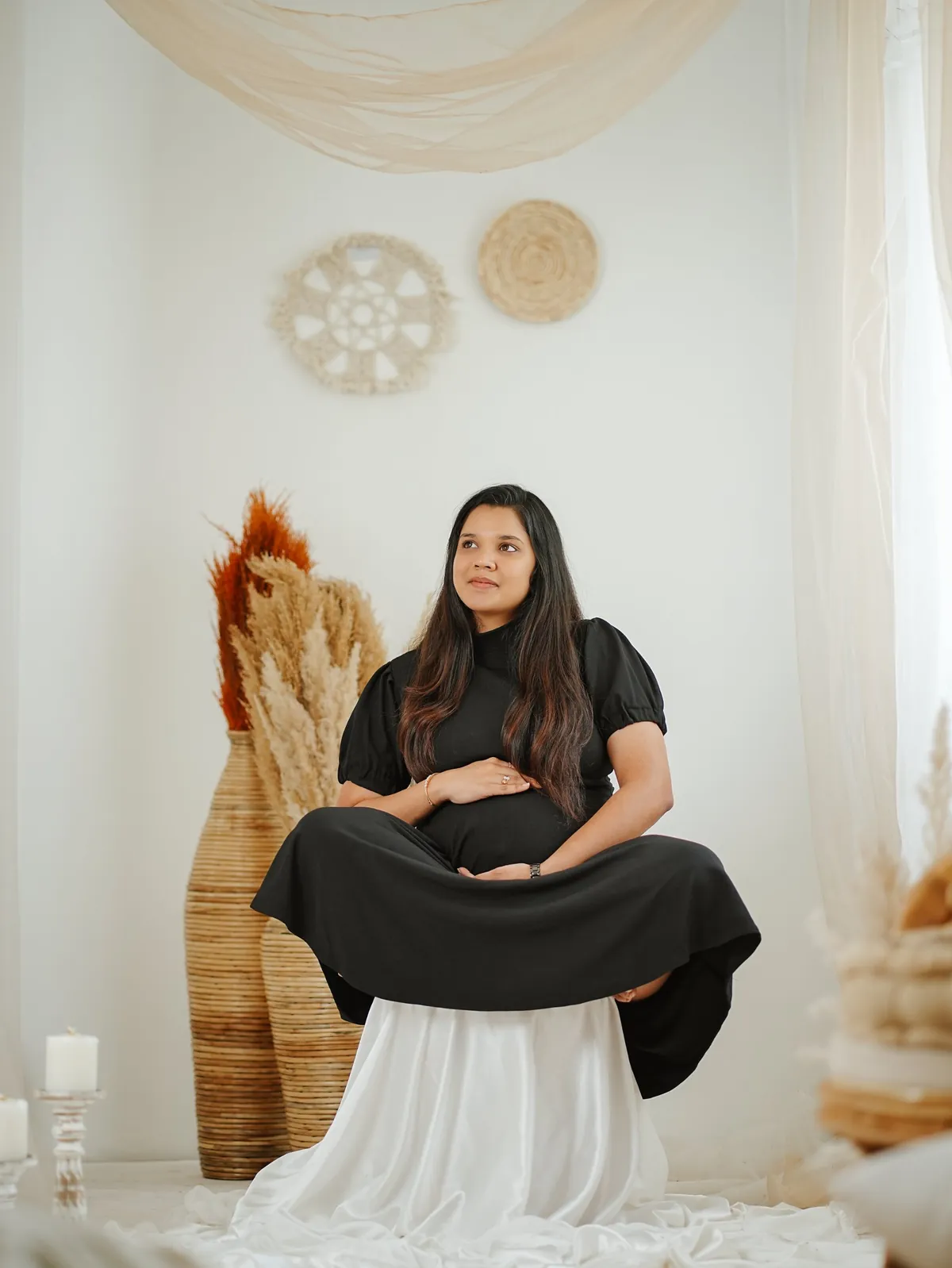 A mother-to-be in a black dress sits cross-legged on a draped stool, hand on her bump, looking away, with pampas grass and candles around her.