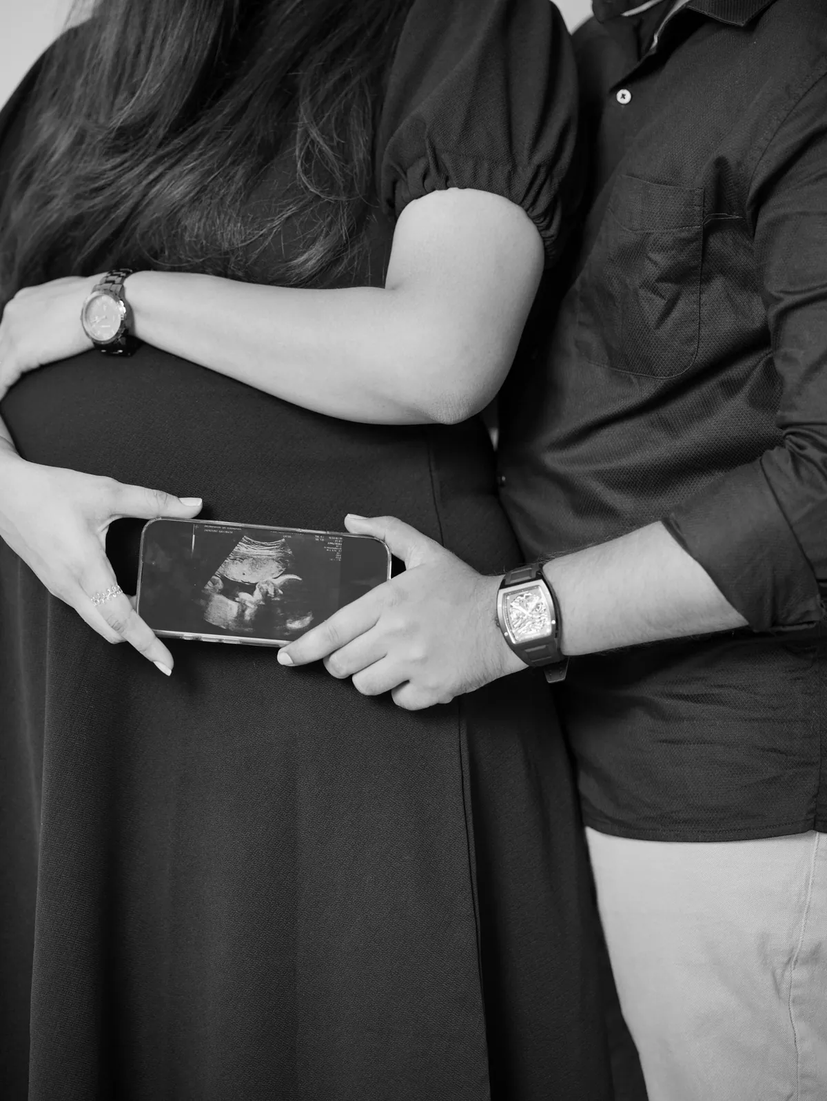 Black and white close-up of a couple in black holding a phone showing an ultrasound scan in front of her bump.