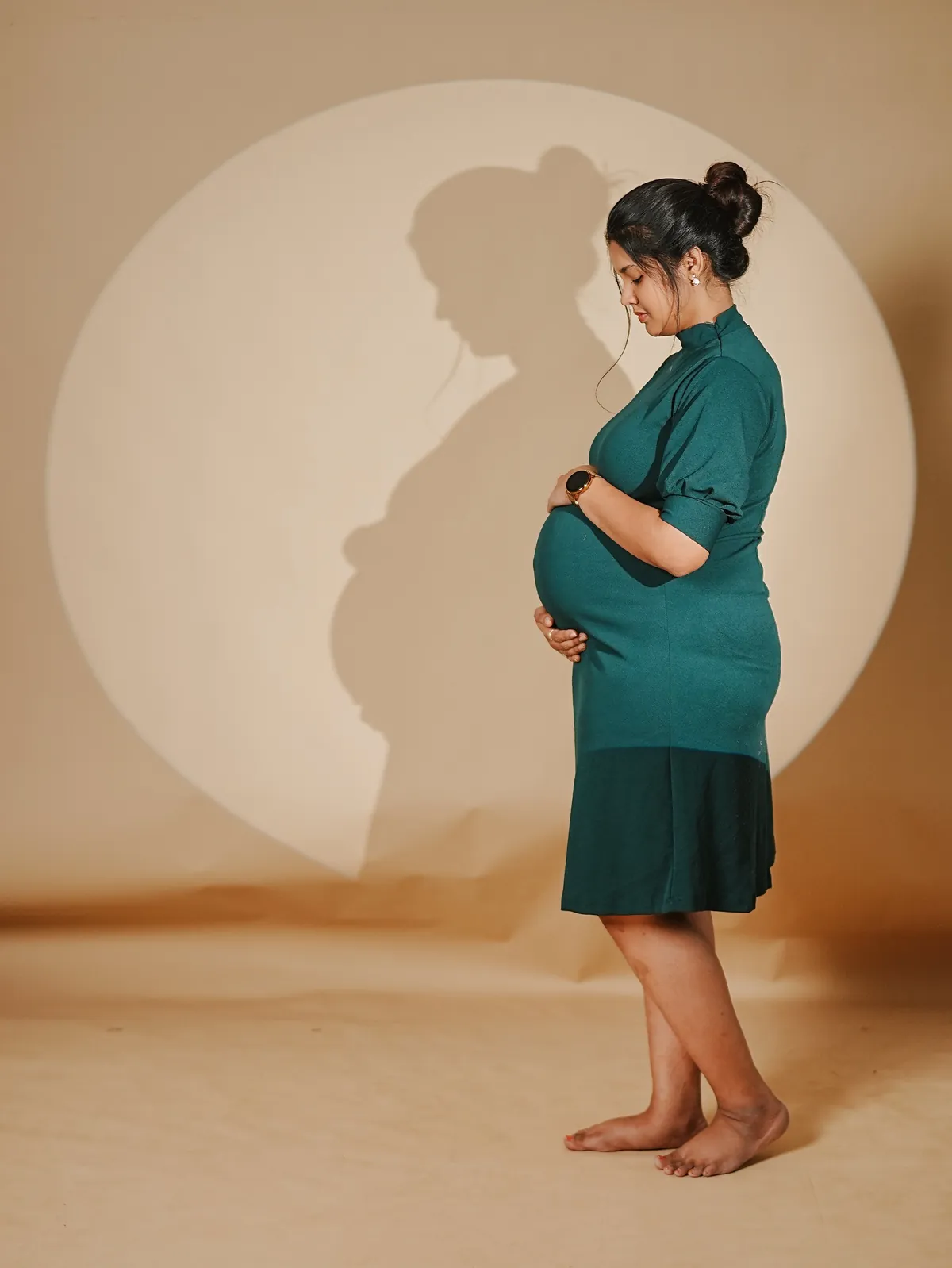 A mother-to-be in a teal dress stands in profile holding her bump, her shadow cast inside a circle of light on a beige backdrop.