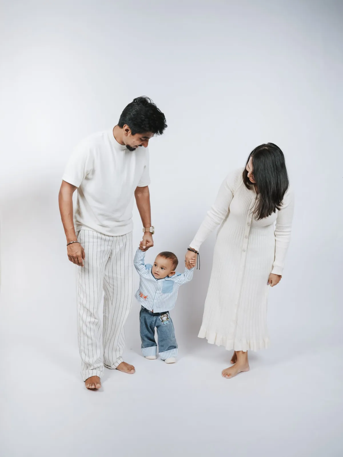 Parents in white each hold a hand of their toddler, who stands between them on a white backdrop.
