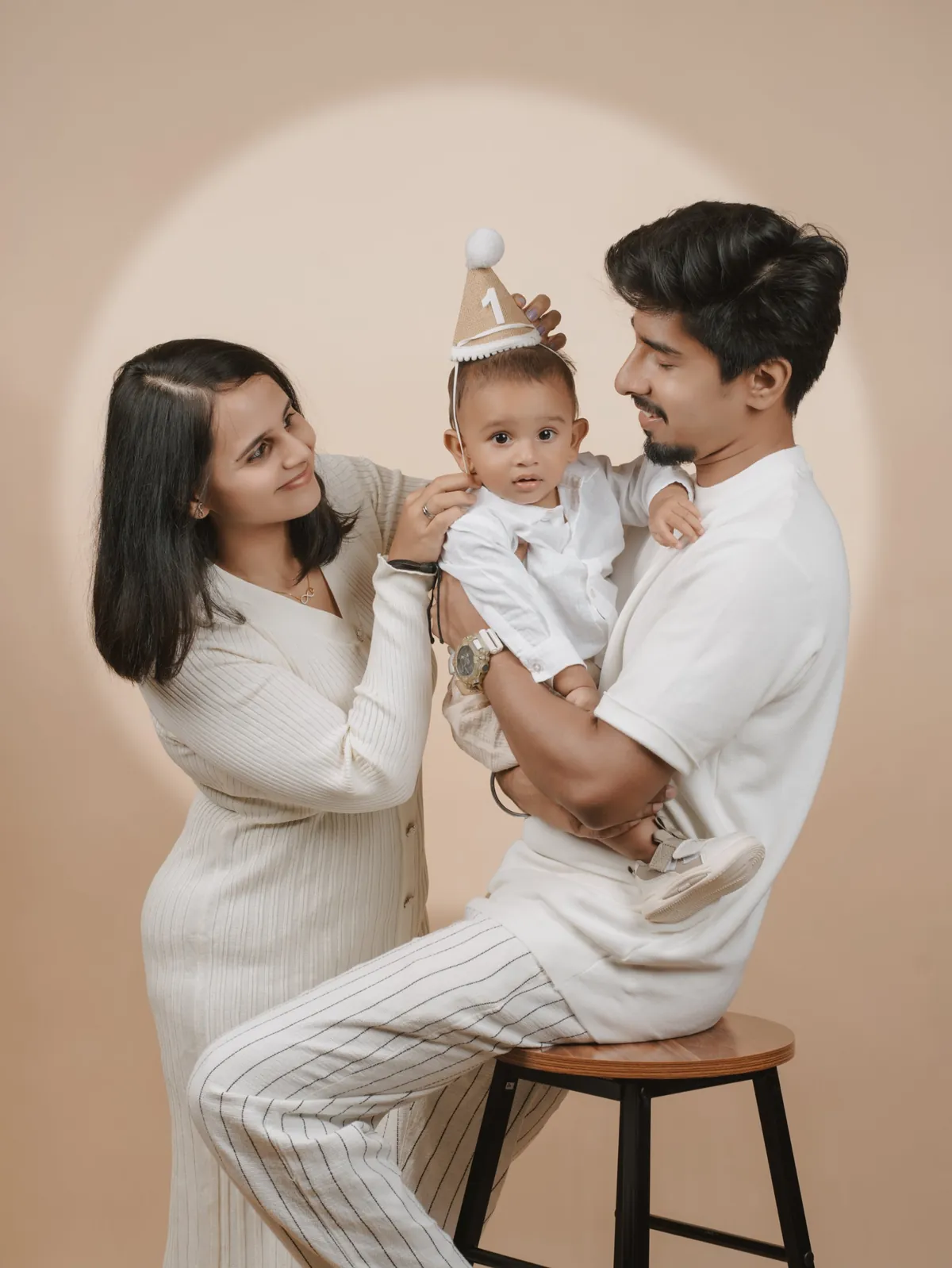 Parents look at their baby, who wears a party hat with the number one and looks at the camera, lit by a soft circle of light on a beige backdrop.
