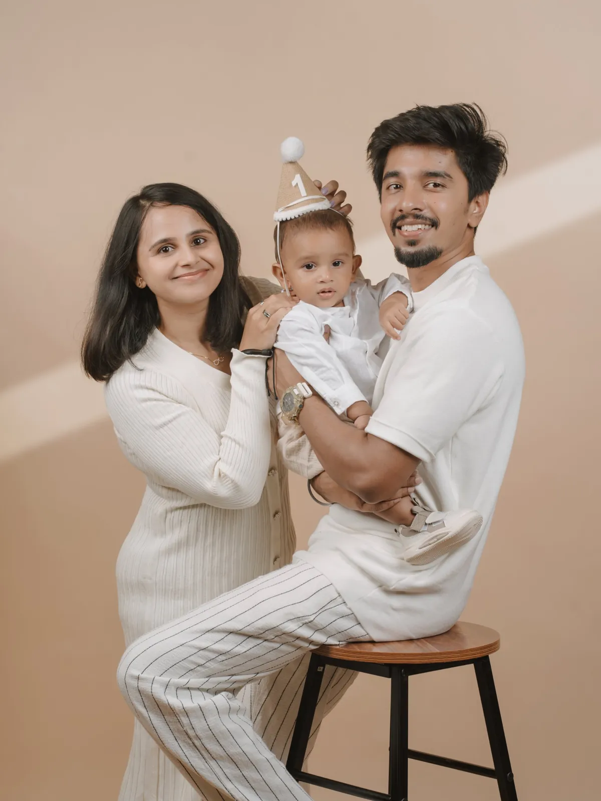 Smiling parents in white hold their baby in a first-birthday party hat against a beige backdrop with a band of light.