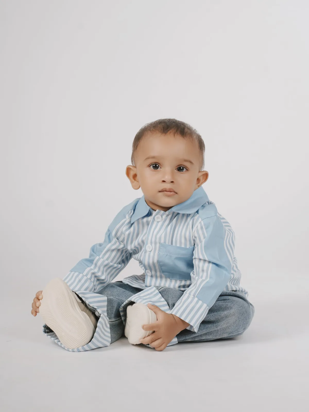 A baby in a blue striped shirt and jeans sits on a white backdrop holding his shoes and looking at the camera.