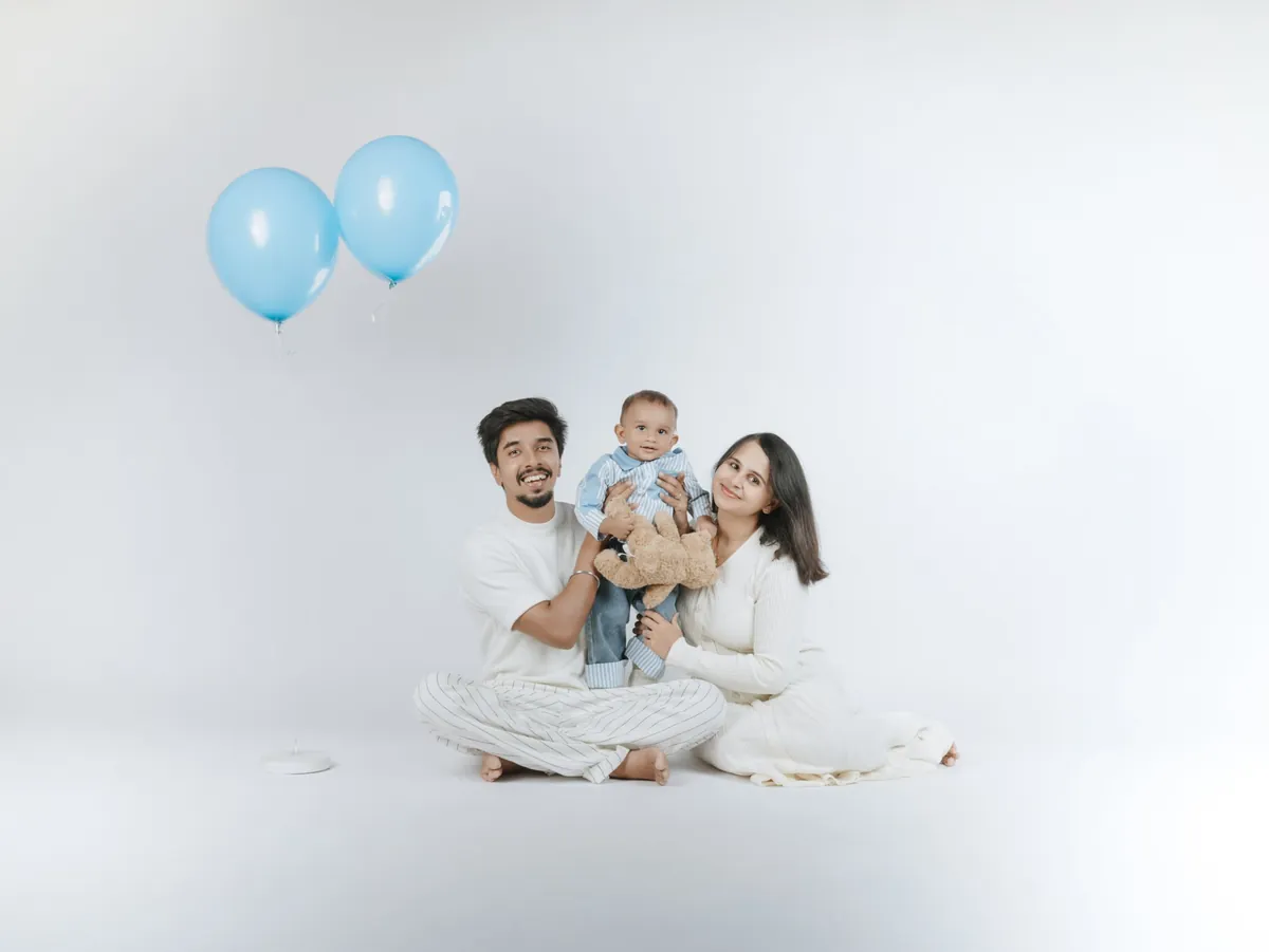 Parents sit on a white studio floor holding their baby and a teddy bear, with two blue balloons floating beside them.