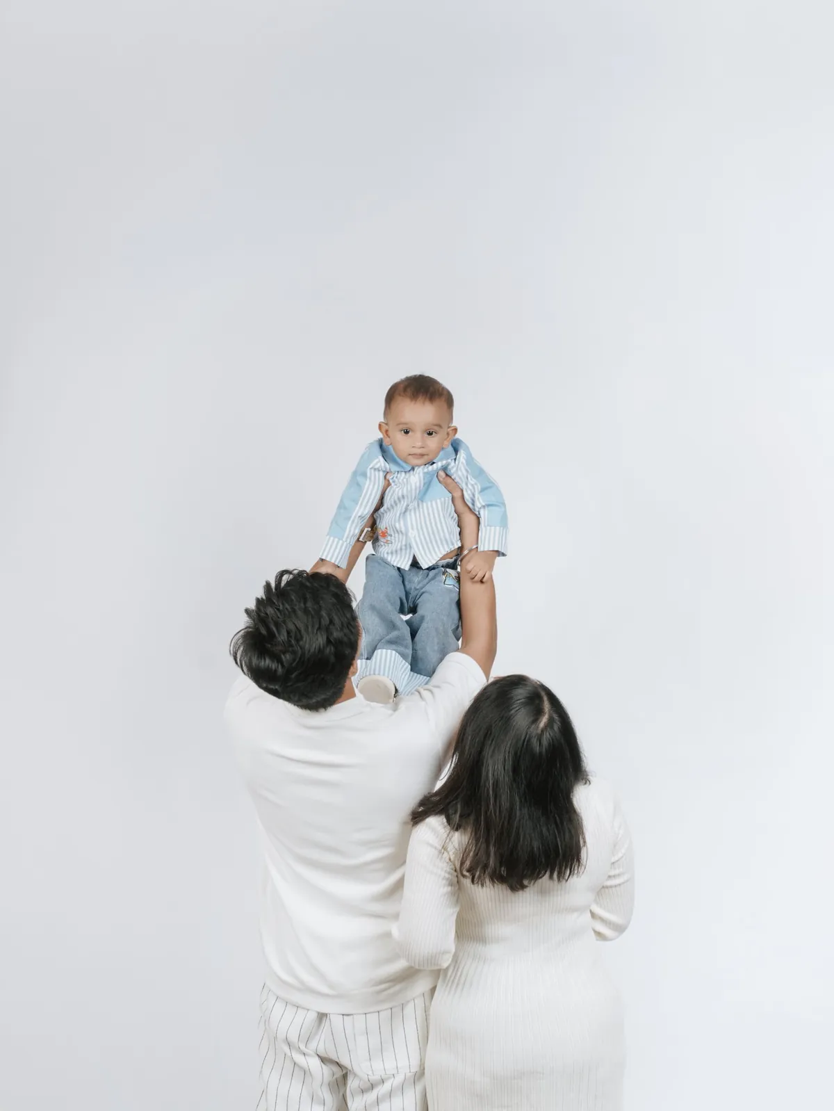 Seen from behind, parents lift their baby high; the baby looks over their heads at the camera on a white backdrop.