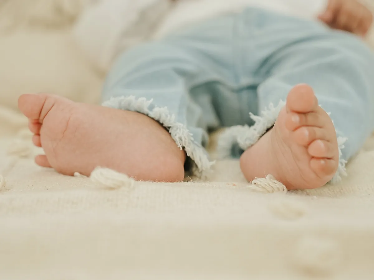 Close view of a baby's bare feet and pale blue trousers on a cream knitted blanket.