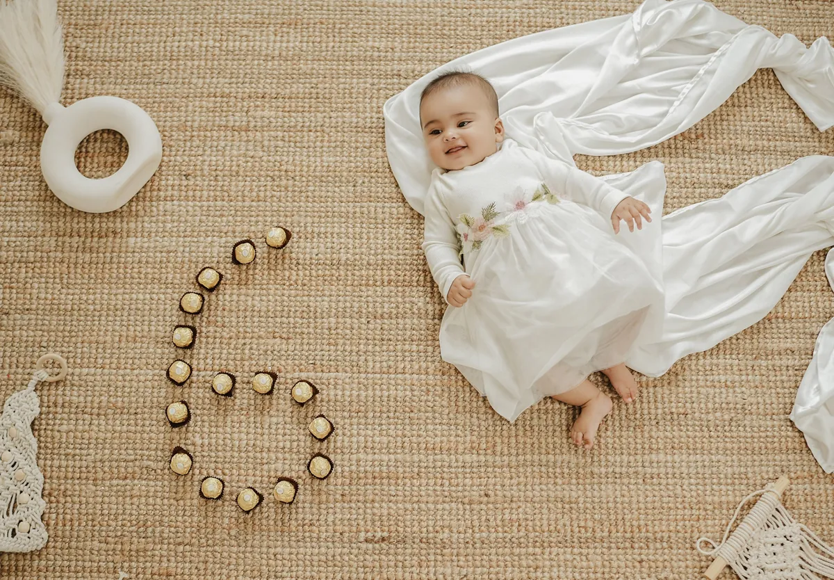 Seen from above, a smiling baby in a white dress lies on a woven mat beside the number six laid out in gold-wrapped chocolates.